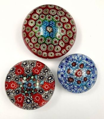 trois presse-papiers en verre, ornés de motifs différents, posés sur un fond blanc