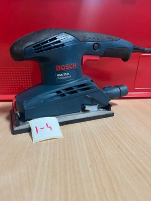 une ponceuse orbitale Bosch posée sur une table en bois, à côté d'une boîte rouge