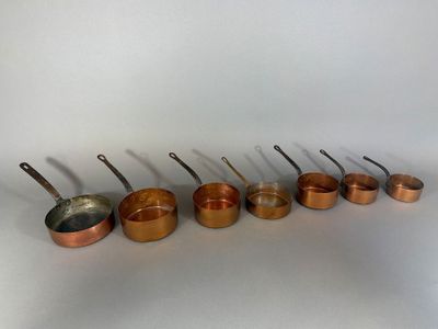 six casseroles et poêles en cuivre alignées sur un plan de travail blanc