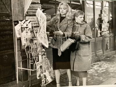 two women standing outside of a store looking at newspapers