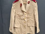 une veste beige aux bordures rouges, suspendue à un cintre devant un mur gris