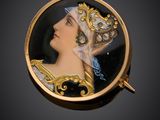 Une broche en or représente une femme portant un chapeau et une écharpe bleue.