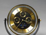 une horloge dorée et noire posée sur un socle, sur une surface blanche