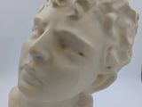 une sculpture blanche représentant un homme aux cheveux bouclés, posée sur une surface blanche