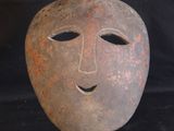 un masque en argile posé sur un socle en bois, sur fond noir