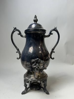 une cafetière ancienne en métal argenté, posée sur un socle et munie d'un couvercle