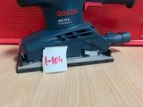 une ponceuse orbitale Bosch posée sur une table, à côté d'une boîte rouge
