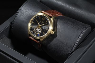 La montre est présentée dans un coffret noir, avec un bracelet en cuir marron.