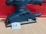 une ponceuse orbitale Bosch posée sur une table, à côté d'une boîte rouge