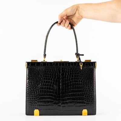 une main tenant un sac à main en cuir de crocodile noir muni de anses dorées