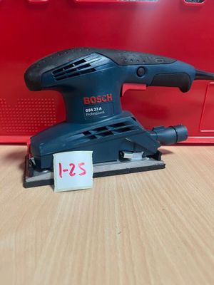une ponceuse orbitale Bosch posée sur une table, avec une boîte rouge en arrière-plan