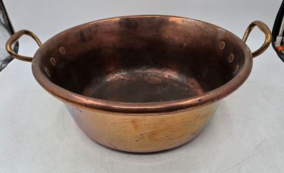 une grande casserole en cuivre avec des poignées, posée sur une table blanche