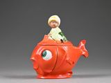 une figurine représentant un garçon assis sur une théière en forme de poisson