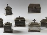 six boîtes en métal argenté, surmontées de croix, posées sur une surface blanche