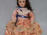 une poupée ancienne assise sur une surface blanche, vêtue d'une robe rose et d'un ruban bleu