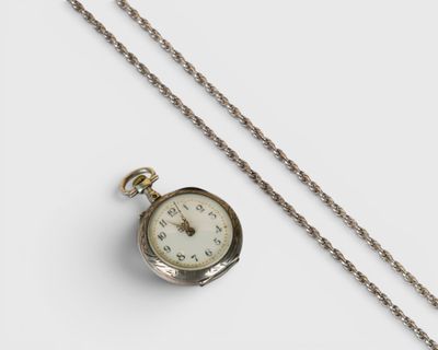une montre de poche en argent, suspendue à une chaîne, posée sur une surface blanche