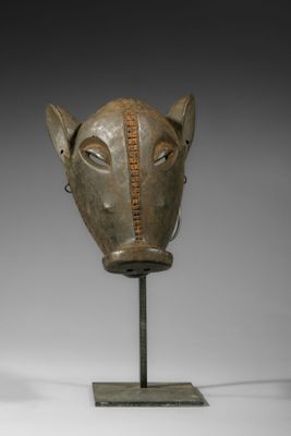 une photo représentant un masque en bois posé sur un socle, sur une surface blanche