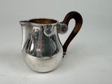 une cruche ancienne en argent avec une poignée en bois, posée sur une surface blanche