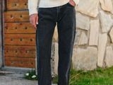 un homme debout devant un mur de pierre, vêtu d'un pull blanc et d'un jean noir