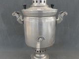 une théière ancienne plaquée argent, posée sur un support, sur un fond gris