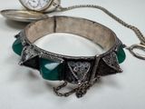 a silver bracelet with green stones on it and a pocket watch in the background