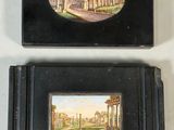 two small black boxes with paintings on them sitting on a white surface