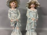 deux figurines représentant deux enfants portant des chapeaux et tenant des fleurs