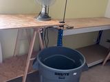 a desk with a fan and a trash can in the corner