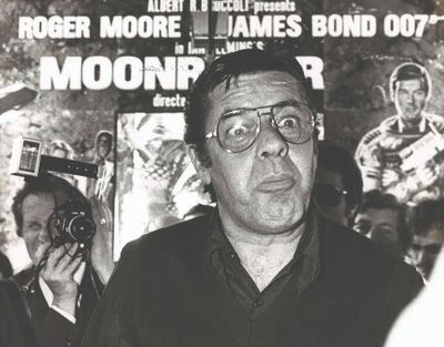 photographie en noir et blanc d'un homme à lunettes debout devant une affiche de cinéma