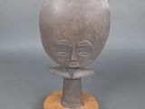 sculpture représentant un visage, posée sur un socle en bois, sur une surface grise