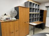 a room with a desk, bookshelf, and a fan in it
