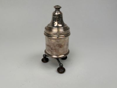 une salière ancienne en métal argenté, montée sur roulettes, posée sur un support blanc