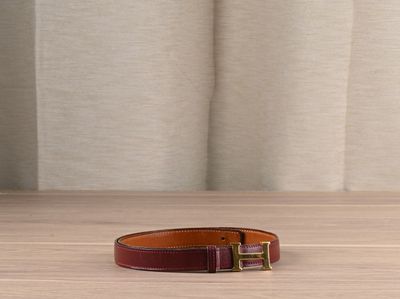 une ceinture en cuir marron avec une boucle dorée, posée sur une table en bois devant un rideau