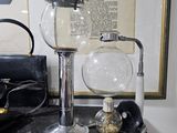 une cafetière en verre posée sur une table, à côté de deux verres et d'une paire de ciseaux