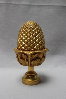 une pomme de pin dorée posée sur un socle, sur fond blanc