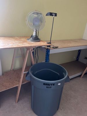 a desk with a fan and a trash can in the corner