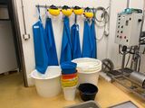 Plusieurs seaux sont alignés dans une cuisine où des tabliers bleus sont accrochés au mur