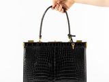 une main tenant un sac à main en cuir de crocodile noir muni de anses dorées