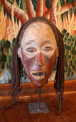 un masque en bois avec des dreadlocks, posé sur un socle devant un tableau