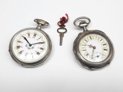 deux montres de poche en argent et une clé posées sur une surface blanche