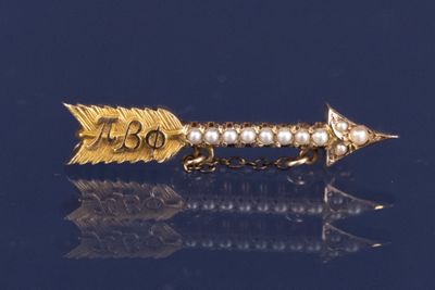 une broche en forme de flèche, en or et perles, sur un fond bleu