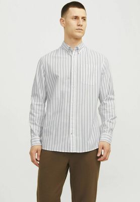Un homme vêtu d'une chemise blanche et d'un pantalon marron se tient debout devant un fond blanc.