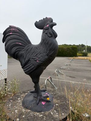 une statue représentant un coq posé sur un rocher dans un parking