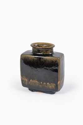 un vase noir avec un couvercle marron posé sur une surface blanche