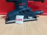 une ponceuse orbitale Bosch posée sur une table, à côté d'une boîte rouge