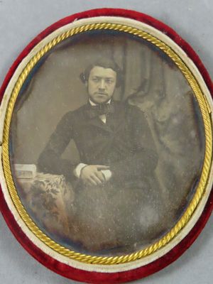 portrait d'un homme en costume noir et nœud papillon, assis sur une chaise
