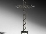 une croix métallique posée sur un socle, dont l'ombre se projette sur le sol