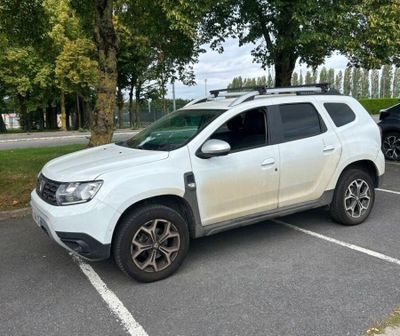 La voiture blanche est garée sur le parking, près des arbres.