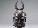 un masque africain muni de cornes, posé sur un socle, sur fond gris