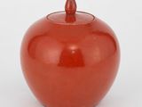 un vase rouge muni d'un couvercle, posé sur une surface blanche